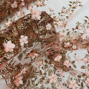 Op de afbeelding: Zachte blush roze bloemen kanten stof met gouden glitter en pailletten. De stof heeft 3D-bloemen met parelcentra en ingewikkelde gouden borduursels, perfect voor bruids- of speciale gelegenheden.
