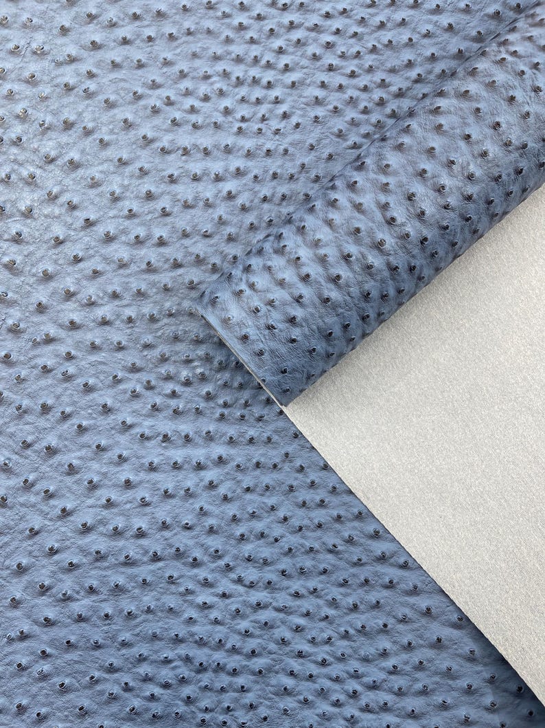 Puede incluir: Primer plano de un rollo de tela de cuero sint&eacute;tico azul con un patr&oacute;n texturizado y punteado. La tela est&aacute; parcialmente enrollada, revelando un reverso blanco. El material tiene un acabado suave y mate, adecuado para varios proyectos de artesan&iacute;a.