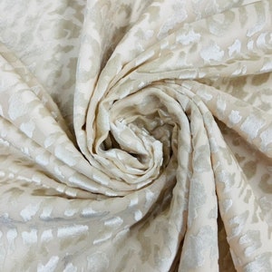 Puede incluir: Tela de color crema con un patrón en relieve de formas abstractas. La tela es suave y lujosa, con un brillo sutil.