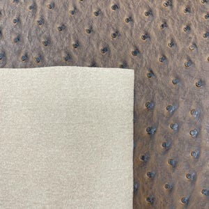 May include: Close-up of brown ostrich leather with a textured surface and small, dark indentations. A section of light beige fabric is visible on the left, contrasting with the leather's pattern. The leather has a pebbled appearance.