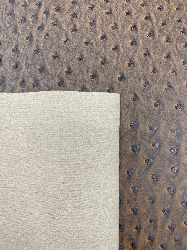 May include: Close-up of brown ostrich leather with a textured surface and small, dark indentations. A section of light beige fabric is visible on the left, contrasting with the leather's pattern. The leather has a pebbled appearance.