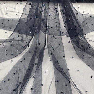 May include: Dark navy blue tulle fabric with scattered, iridescent beads. The sheer material drapes in soft folds, creating a flowing effect. The beads add a subtle shimmer, enhancing the fabric's texture. Ideal for bridal wear, evening gowns, or decorative projects.