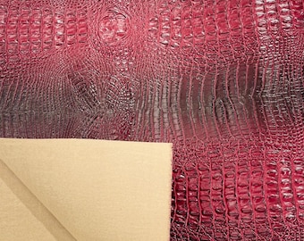 Könnte beinhalten: Nahaufnahme eines dunkelroten Kunstkrokodillederstoffs mit einem subtilen Ombre-Effekt. Der Stoff hat ein strukturiertes, geprägtes Muster, das an Krokodilschuppen erinnert.