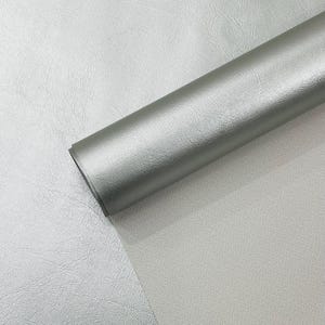 Può includere: Un rotolo di materiale in similpelle grigio argento. Il materiale ha una superficie strutturata ed è parzialmente srotolato, rivelando il lato inferiore. Il colore è una tonalità neutra e attenuata, adatta a vari progetti di artigianato.