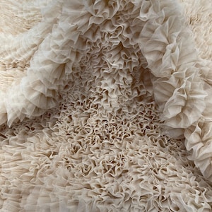 Könnte beinhalten: Nahaufnahme eines beigefarbenen Stoffes mit einer geschichteten, gerüschten Textur. Der Stoff scheint Tüll oder ein ähnliches leichtes Material zu sein, das einen weichen, fließenden Effekt erzeugt. Die Rüschen sind dicht gepackt und verleihen dem Textil Volumen und visuelles Interesse.