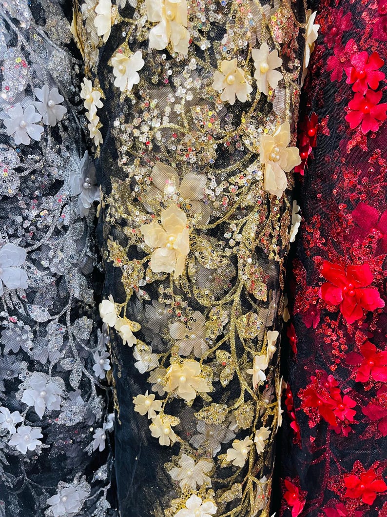 May include: Three rolls of black mesh fabric with different colored 3D floral embellishments. The fabric on the left has silver sequins and white flowers. The middle fabric has gold sequins and cream flowers. The fabric on the right has red flowers.