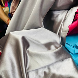 May include: A close-up of a silver satin fabric with a smooth, silky texture. The fabric is folded and draped, creating soft folds and curves.