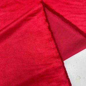 Peut inclure: Gros plan sur un tissu rouge vif, possiblement une couverture ou un plaid, avec une texture douce et veloutée. Le tissu est plié, révélant un envers rouge légèrement plus clair. Les bords sont inachevés, montrant la construction du tissu.