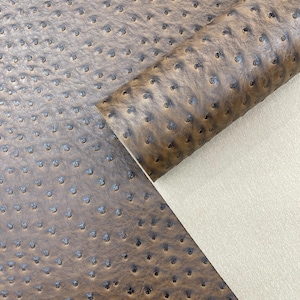 May include: Close-up of brown faux ostrich leather with a textured surface and small, dark indentations. A rolled edge reveals a beige backing, showcasing the material's construction. This fabric is suitable for various crafting projects.
