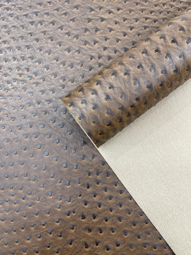 May include: Close-up of brown faux ostrich leather with a textured surface and small, dark indentations. A rolled edge reveals a beige backing, showcasing the material's construction. This fabric is suitable for various crafting projects.