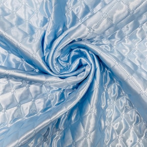 Puede incluir: Tela de satén acolchada azul claro con un patrón de diamantes. La tela está doblada y drapeada, creando un efecto arremolinado. El satén tiene una superficie brillante y reflectante.