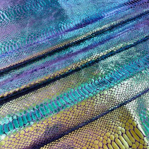 Könnte beinhalten: Nahaufnahme eines schillernden Stoffes mit Schlangenmuster. Der Stoff weist eine Mischung aus Farben auf, darunter Blau, Lila, Türkis und Gold. Das Material ist gefaltet, wodurch Schatten entstehen und die Textur der Schuppen hervorgehoben wird.