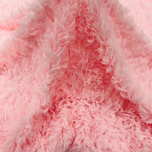 Puede incluir: Primer plano de una tela suave y con volantes en un tono rosa claro. La tela está densamente superpuesta, creando una apariencia texturizada similar a una nube. Las delicadas capas sugieren un material delicado, posiblemente para un vestido o un artículo decorativo.