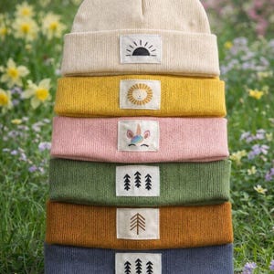 Könnte beinhalten: Ein Stapel von sieben Strickmützen in verschiedenen Farben, darunter Creme, Gelb, Rosa, Grün, Braun und Blau. Jede Mütze hat ein rechteckiges Patch mit einem anderen Design: eine Sonne, ein Löwe, ein Einhorn und Bäume.