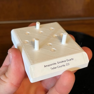 May include: White plastic display stand with three small white posts. The stand has nine holes and a label that reads "Amazonite, Smokey Quartz Teller County, CO".