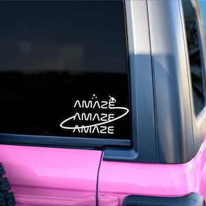 May include: White vinyl decal with the word "AMAZE" in a stylized font, applied to the rear window of a pink vehicle. The decal includes a surfer graphic and a curved line. The background shows a beach scene.