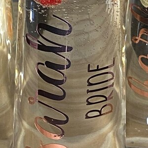 May include: A clear glass champagne flute with a gold bow and the words "Bride to be" written on it. The flute is filled with a clear liquid and has fresh fruit and berries floating inside.