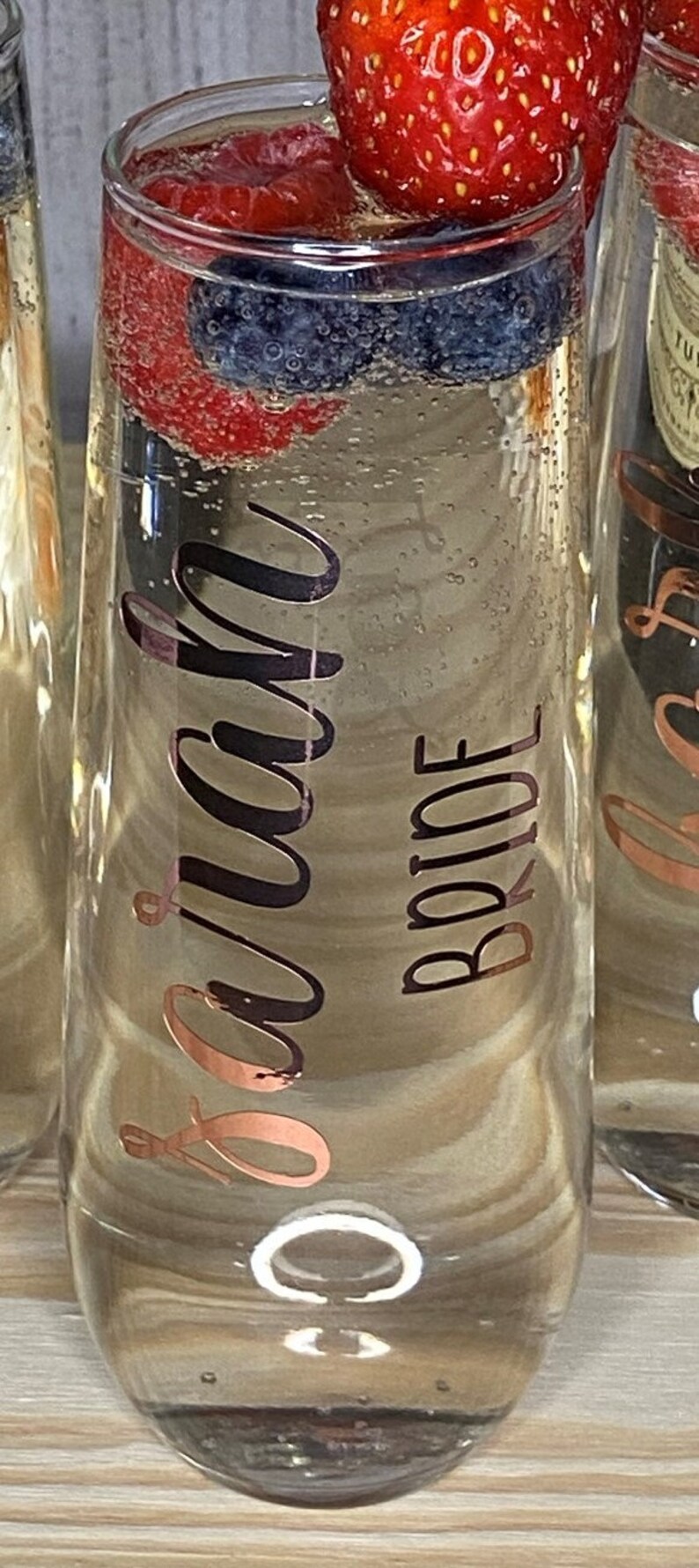 May include: A clear glass champagne flute with a gold bow and the words "Bride to be" written on it. The flute is filled with a clear liquid and has fresh fruit and berries floating inside.