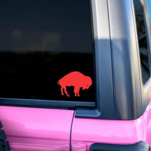 May include: A red buffalo silhouette decal on a pink car window. The car is parked on a beach with palm trees in the background.