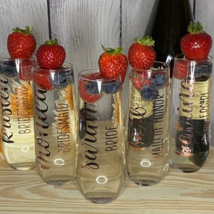 May include: Six champagne flutes with gold lettering. The flutes are filled with a clear liquid and garnished with fresh fruit. The lettering on the flutes reads "Bride", "Maid of Honor", "Bridesmaid", "Bridesmaid", "Bridesmaid", and "Bridesmaid".