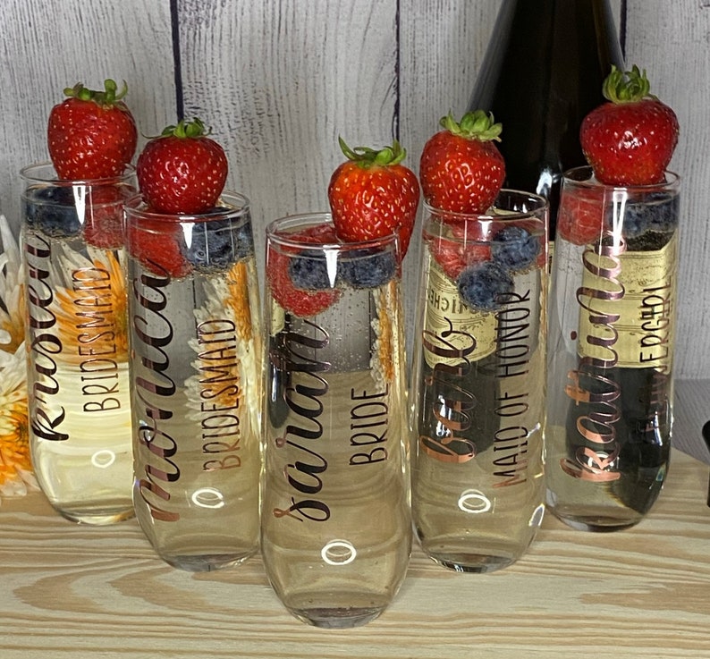 May include: Six champagne flutes with gold lettering. The flutes are filled with a clear liquid and garnished with fresh fruit. The lettering on the flutes reads "Bride", "Maid of Honor", "Bridesmaid", "Bridesmaid", "Bridesmaid", and "Bridesmaid".