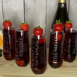 May include: Six champagne flutes with different titles, including "Bride", "Maid of Honor", "Bridesmaid", and "Flower Girl". Each flute is decorated with a strawberry and filled with a red beverage.