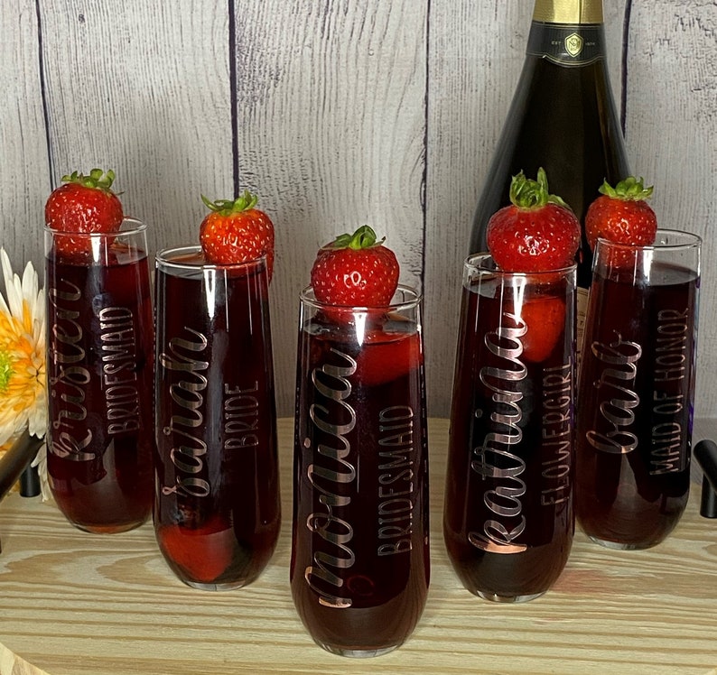 May include: Six champagne flutes with different titles, including "Bride", "Maid of Honor", "Bridesmaid", and "Flower Girl". Each flute is decorated with a strawberry and filled with a red beverage.