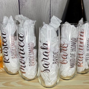 May include: Six champagne flutes with rose gold lettering. The flutes are labeled "Kristen Bridesmaid", "Monica Bridesmaid", "Sarah Bride", "Barb Maid of Honor", "Lexi", and "Flowergirl".