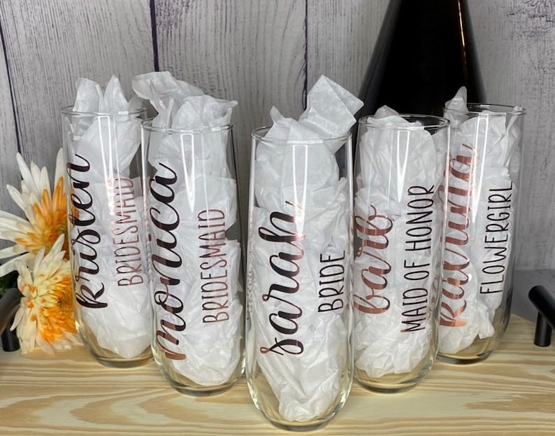 May include: Six champagne flutes with rose gold lettering. The flutes are labeled "Kristen Bridesmaid", "Monica Bridesmaid", "Sarah Bride", "Barb Maid of Honor", "Lexi", and "Flowergirl".
