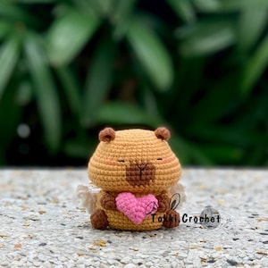 Könnte beinhalten: Ein gehäkeltes braunes und hellbraunes Tier, das ein rosa Herz hält. Das Tier hat einen kleinen, runden Körper und ein süßes Gesicht. Das Tier sitzt auf einer grauen Oberfläche. Der Text "Tokki Crochet" ist in der unteren rechten Ecke des Bildes sichtbar.