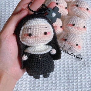 May include: A crocheted nun keychain with a black dress, white head covering, and a silver cross. The keychain has a black flower charm and a silver ring.
