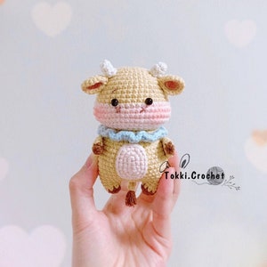 May include: A small crocheted cow amigurumi toy with a light brown body, pink cheeks, and a blue and white ruffle collar. The toy is being held in a hand against a light blue background with white hearts.