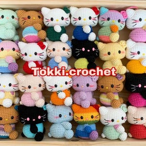 May include: A collection of 20 crocheted amigurumi toys in various colors and designs. The toys are all small and feature different animal characters, including cats, bears, and bunnies. The toys are arranged in a wooden box with the text "Tokki.crochet" printed on the front.