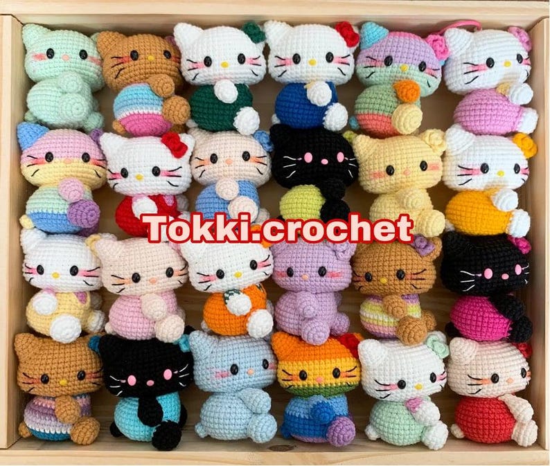 May include: A collection of 20 crocheted amigurumi toys in various colors and designs. The toys are all small and feature different animal characters, including cats, bears, and bunnies. The toys are arranged in a wooden box with the text "Tokki.crochet" printed on the front.