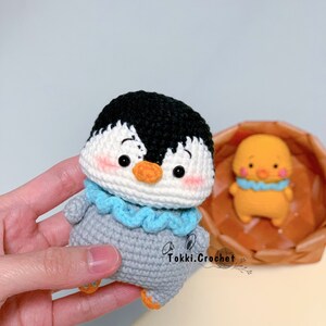 May include: A crocheted penguin toy with a black head, white face, gray body, and blue scarf. The penguin has a small orange beak and black eyes. The toy is being held in a hand.