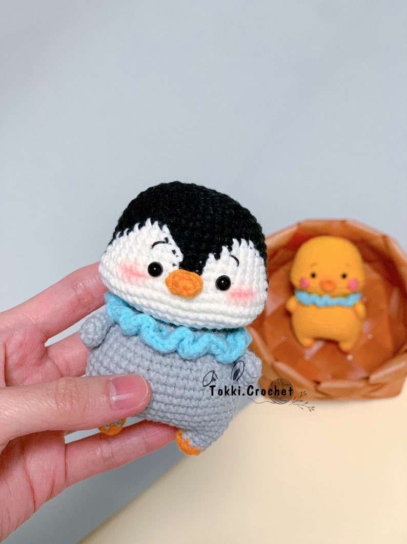 May include: A crocheted penguin toy with a black head, white face, gray body, and blue scarf. The penguin has a small orange beak and black eyes. The toy is being held in a hand.
