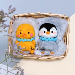 May include: Two crocheted stuffed animals, a yellow chick and a gray penguin, are sitting in a wicker basket lined with white tulle. Both animals have blue crocheted collars and embroidered faces.