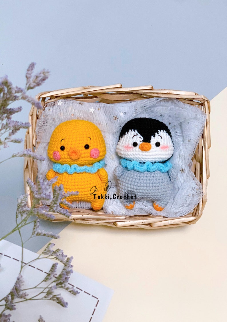 May include: Two crocheted stuffed animals, a yellow chick and a gray penguin, are sitting in a wicker basket lined with white tulle. Both animals have blue crocheted collars and embroidered faces.