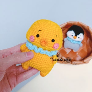 May include: A yellow crocheted chick with blue and pink accents. The chick has a small beak and large eyes. The chick is holding a blue crocheted scarf.