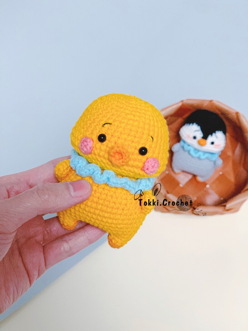 May include: A yellow crocheted chick with blue and pink accents. The chick has a small beak and large eyes. The chick is holding a blue crocheted scarf.