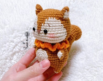 Könnte beinhalten: Ein gehäkeltes Eichhörnchen-Amigurumi-Spielzeug mit braunem und weißem Fell, das einen orangefarbenen Rüschenkragen trägt. Das Spielzeug wird in einer Hand vor einem weißen, flauschigen Hintergrund gehalten.