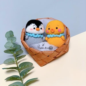 May include: A crocheted penguin and chick in a woven basket. The penguin is gray with a white belly and black eyes. The chick is yellow with a blue beak and black eyes. The basket is brown and has a white label that says "Tokki. Crochet".