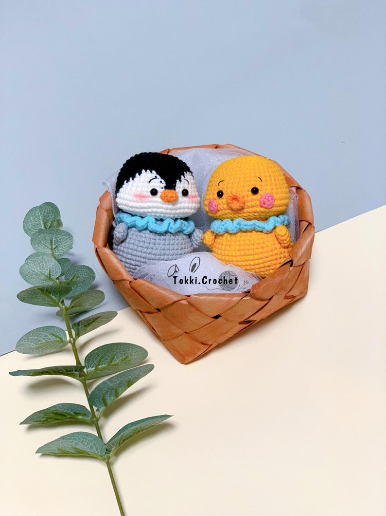 May include: A crocheted penguin and chick in a woven basket. The penguin is gray with a white belly and black eyes. The chick is yellow with a blue beak and black eyes. The basket is brown and has a white label that says "Tokki. Crochet".