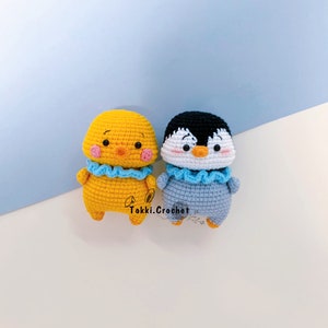 May include: Two crocheted stuffed animals, a yellow chick and a gray penguin, both wearing blue ruffled collars. The chick has a pink face and the penguin has a white face. The animals are sitting on a white surface.