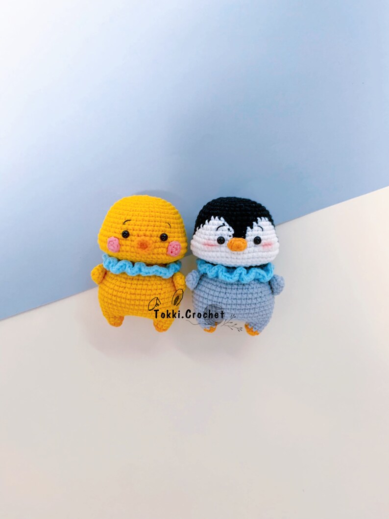May include: Two crocheted stuffed animals, a yellow chick and a gray penguin, both wearing blue ruffled collars. The chick has a pink face and the penguin has a white face. The animals are sitting on a white surface.
