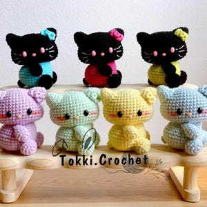 May include: Seven crocheted cat plushies in various colors, including purple, green, yellow, and blue, are displayed on a wooden shelf. The plushies are arranged in a row, with the black cat plushies at the front of the shelf. The shelf has the text "Tokki.Crochet" printed on it.