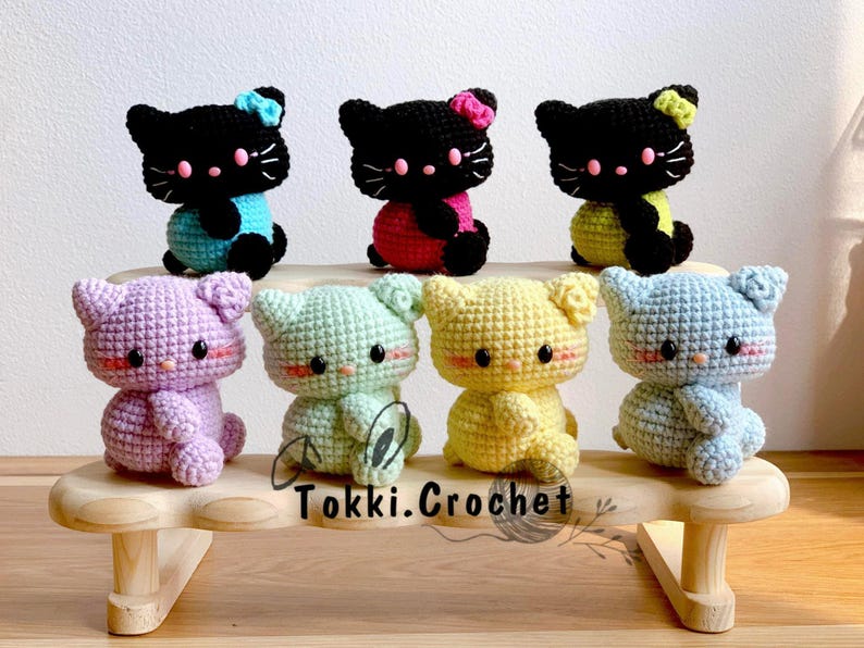May include: Seven crocheted cat plushies in various colors, including purple, green, yellow, and blue, are displayed on a wooden shelf. The plushies are arranged in a row, with the black cat plushies at the front of the shelf. The shelf has the text "Tokki.Crochet" printed on it.