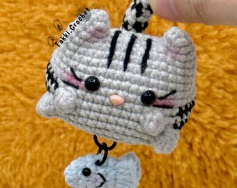 May include: Crocheted gray cat with black stripes and pink accents, holding a small blue fish. The cat has black button eyes and a pink nose. The cat's tail is black and white striped. The cat is attached to a black loop.