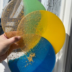 May include: Three round resin coasters with a layered design. The top coaster is green and gold, the middle coaster is yellow and gold, and the bottom coaster is blue and gold.