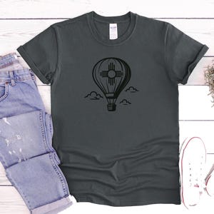 May include: Dark gray t-shirt featuring a black hot air balloon design with the New Mexico Zia symbol. The shirt is paired with blue jeans and white sneakers, suggesting a casual, everyday style. The design includes clouds.